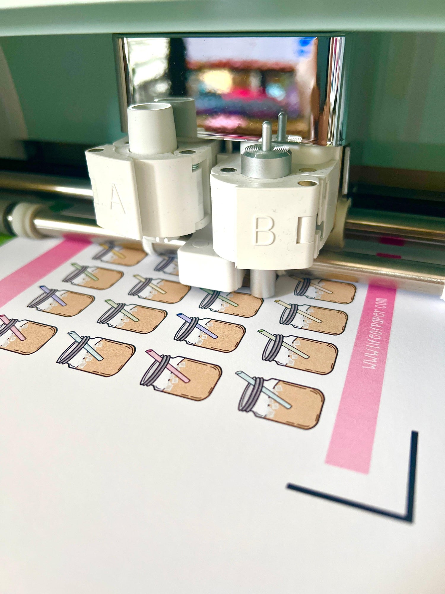 Close-up of a cutting machine working on a sheet of paper with multiple illustrations of mason jar drinks on it. The illustrations are arranged in rows, each featuring a mason jar with a straw.