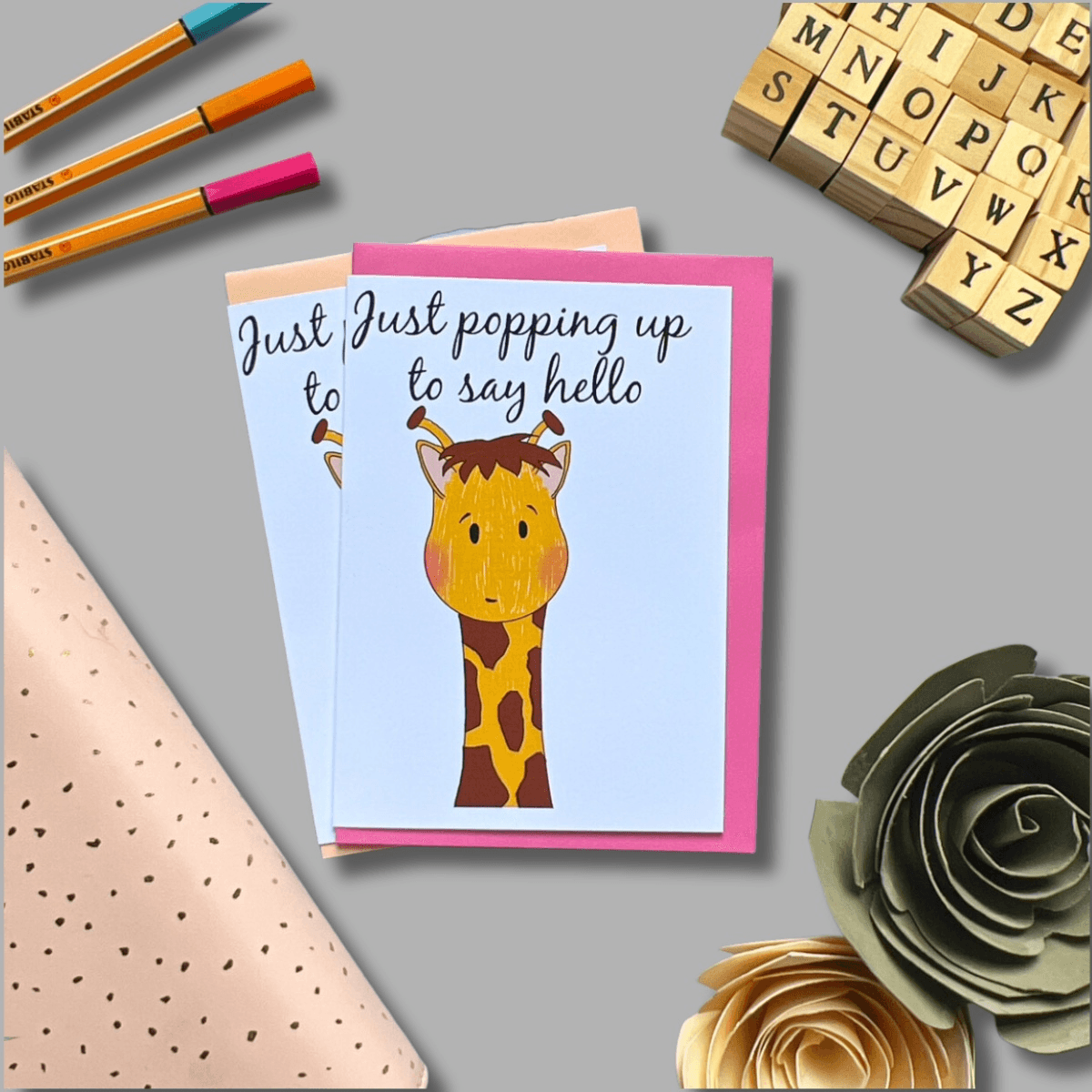 The "Poppin' up to say Hello" card, featuring an adorable giraffe illustration and the text "Just popping up to say hello," is surrounded by colored pencils, a block letter stamp set, and paper flowers in pink and grey tones. Another card lies beneath it, slightly hidden—perfect for friends or sending a note.