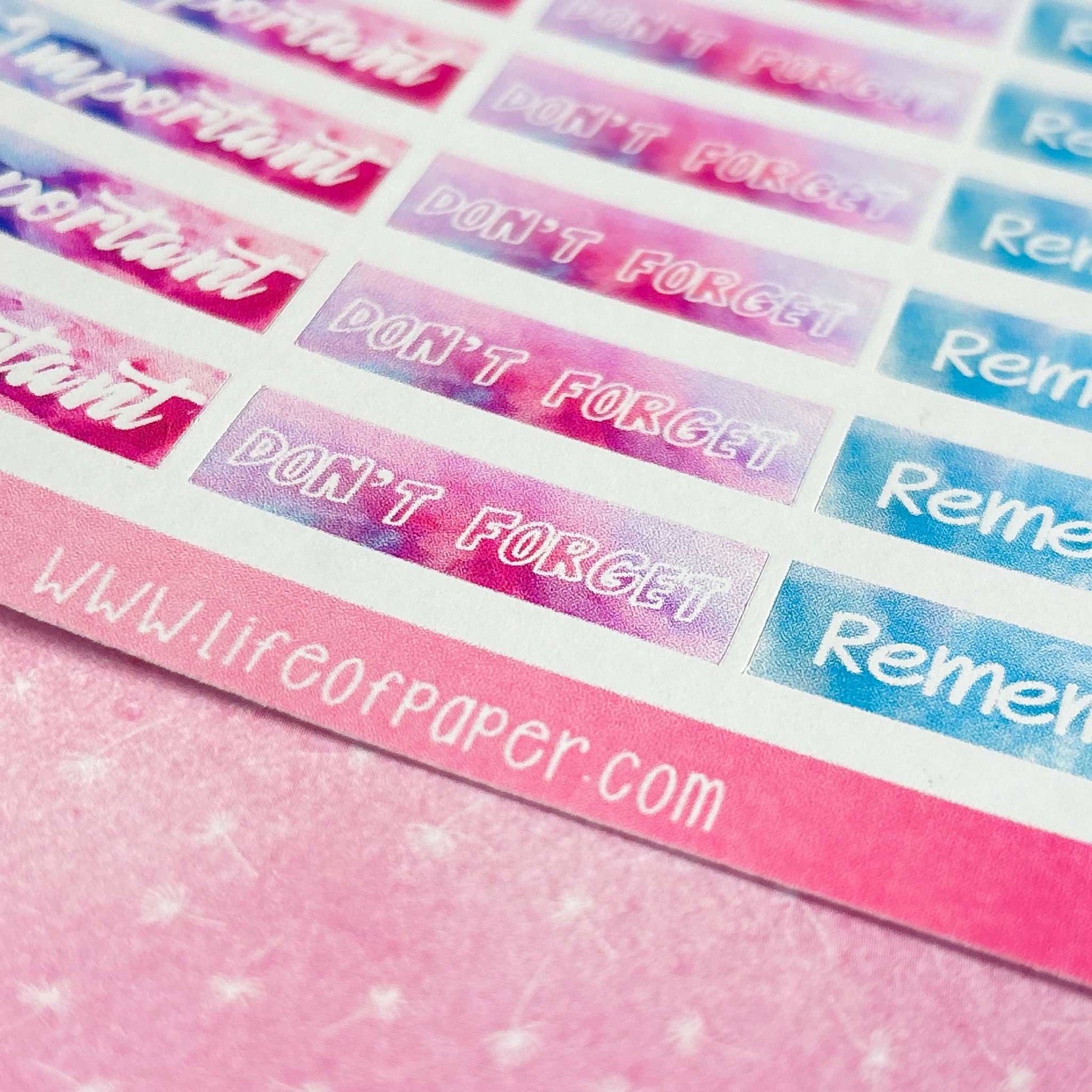 Close-up of colorful Banner Stickers on a pink surface, featuring the phrases "DON'T FORGET" and "Remember" as reminder headings. The website "www.lifeofpaper.com" appears at the bottom. Perfect to help you organize your planner with style.