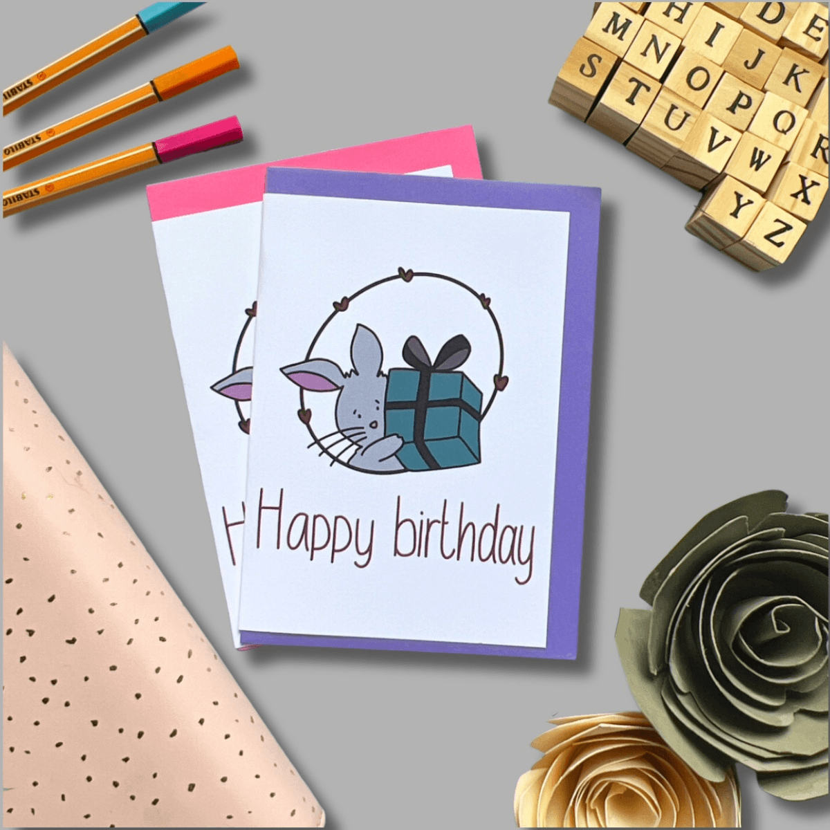 A flat lay photo showcasing two "Cute Bunny Birthday Cards" with a delightful mouse illustration and the text "Happy birthday" on the front. Accompanied by three pens, a sheet of paper adorned with black dots, charming sweet bunny decorations, and wooden alphabet stamps, all set against a gray background.