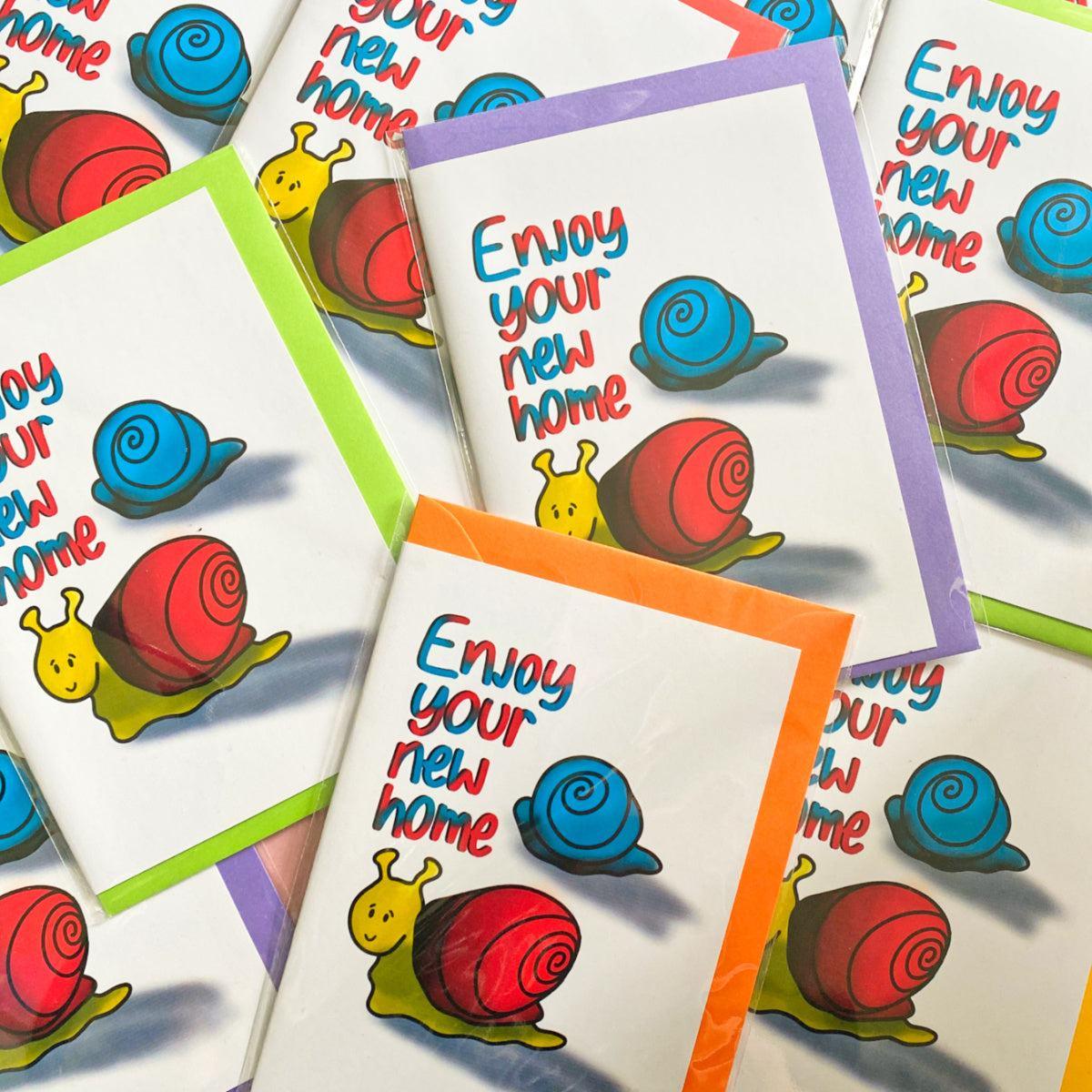 The New Home Greeting Card showcases a joyful assortment of vibrant cards, each depicting a whimsical cartoon snail with a yellow body and either a red or blue shell. The cheerful message "Enjoy your new home" is presented in an engaging, handwritten-style font. These delightful snail-themed greeting cards come adorned with assorted colored borders, including green, orange, and purple, all packaged in eco-friendly biodegradable materials.