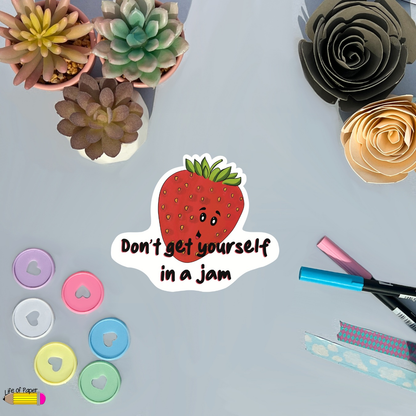 A flat lay of stationery items surrounds a strawberry sticker with a frowning face and the text "Don't get yourself in a jam." The scene features succulent plants, markers, heart-shaped magnets, rolls of tape, and the charming Don't get in a Jam Vinyl sticker.