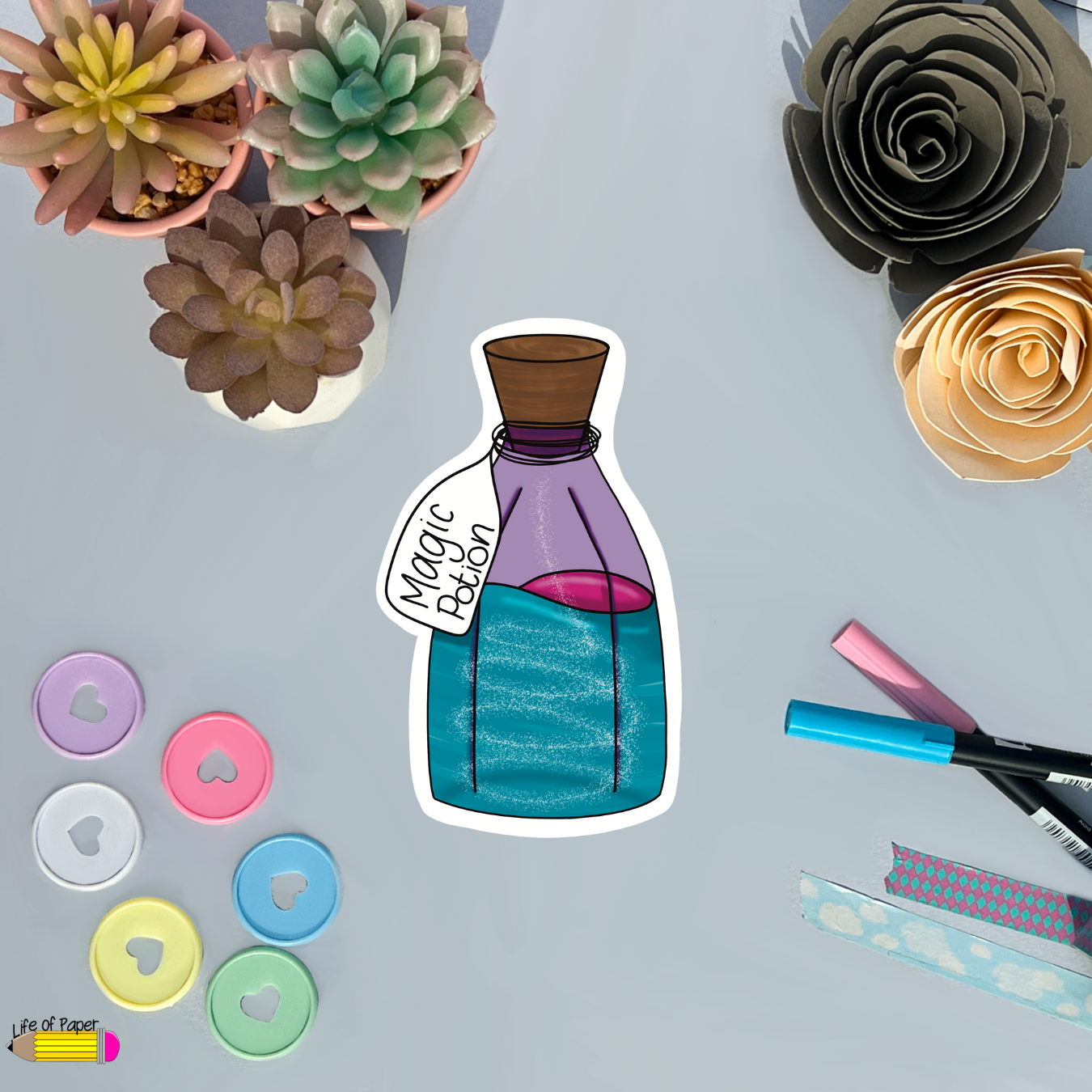A Magic Potion Vinyl Sticker is centered on a table. Surrounding the Magic Potion Vinyl Sticker are various items: colorful succulent plants, pastel buttons, pink and blue markers, and decorative paper roses. The strong matte vinyl adds an extra touch of durability and charm.