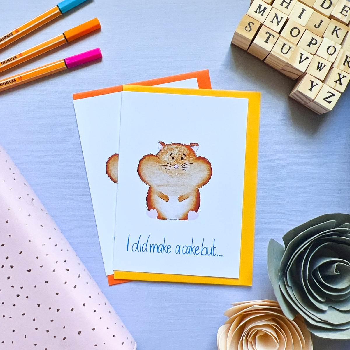 A flat lay of the Hamster Greeting Card, featuring an illustration of a hamster holding a cake. The card reads, "I did make a cake but..." The background includes orange pens, wooden letter blocks, a pink polka-dotted cloth, and paper flowers. This charming birthday card is perfect for any celebration.