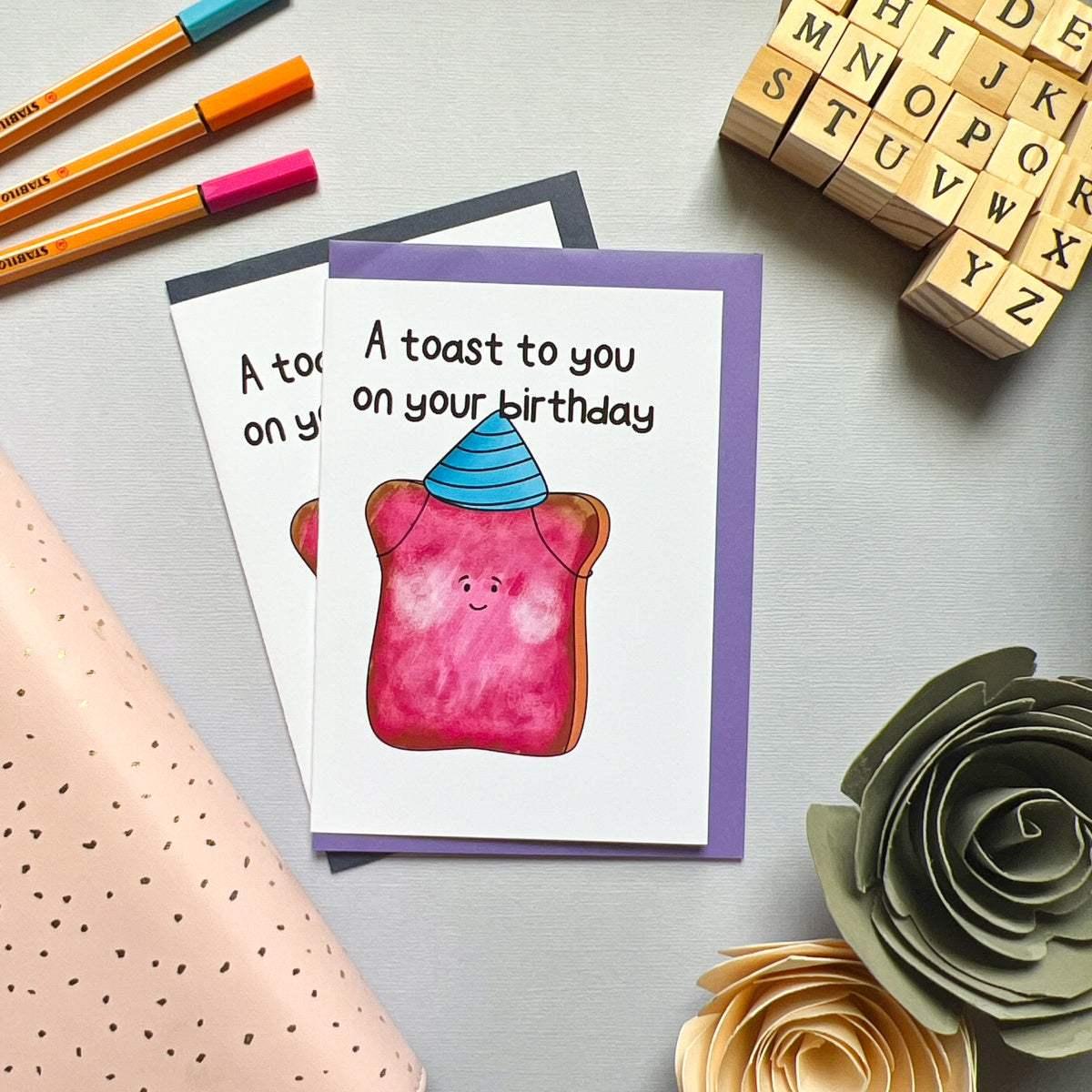 The Birthday Toast Birthday Card features an illustration of a slice of toast wearing a blue party hat and smiling. The card reads, "A toast to you on your special day." It is displayed on a table alongside another identical card, surrounded by pens, letter stamps, and paper flowers.