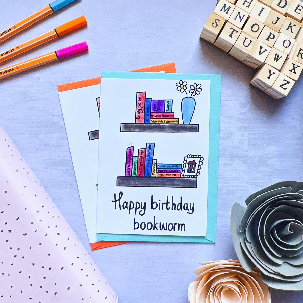 The Bookworm Birthday Card, adorned with a cheerful "Happy birthday bookworm" message, showcases two illustrated bookshelves brimming with books, a vase of flowers, and a mug. Surrounding the card are other delightful cards, vibrant pens, polka-dot gift wrap, hand-crafted flowers, and charming wooden letter blocks.