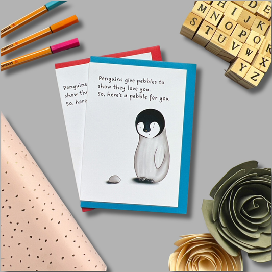 A desk featuring two "Penguin card" greeting cards with an image of a penguin giving a pebble, surrounded by colored pens, a wooden letter stamp set, biodegradable polka-dotted wrapping paper, and paper-crafted roses. The card text reads: "Penguins give pebbles to show love. So, here's a pebble for you.