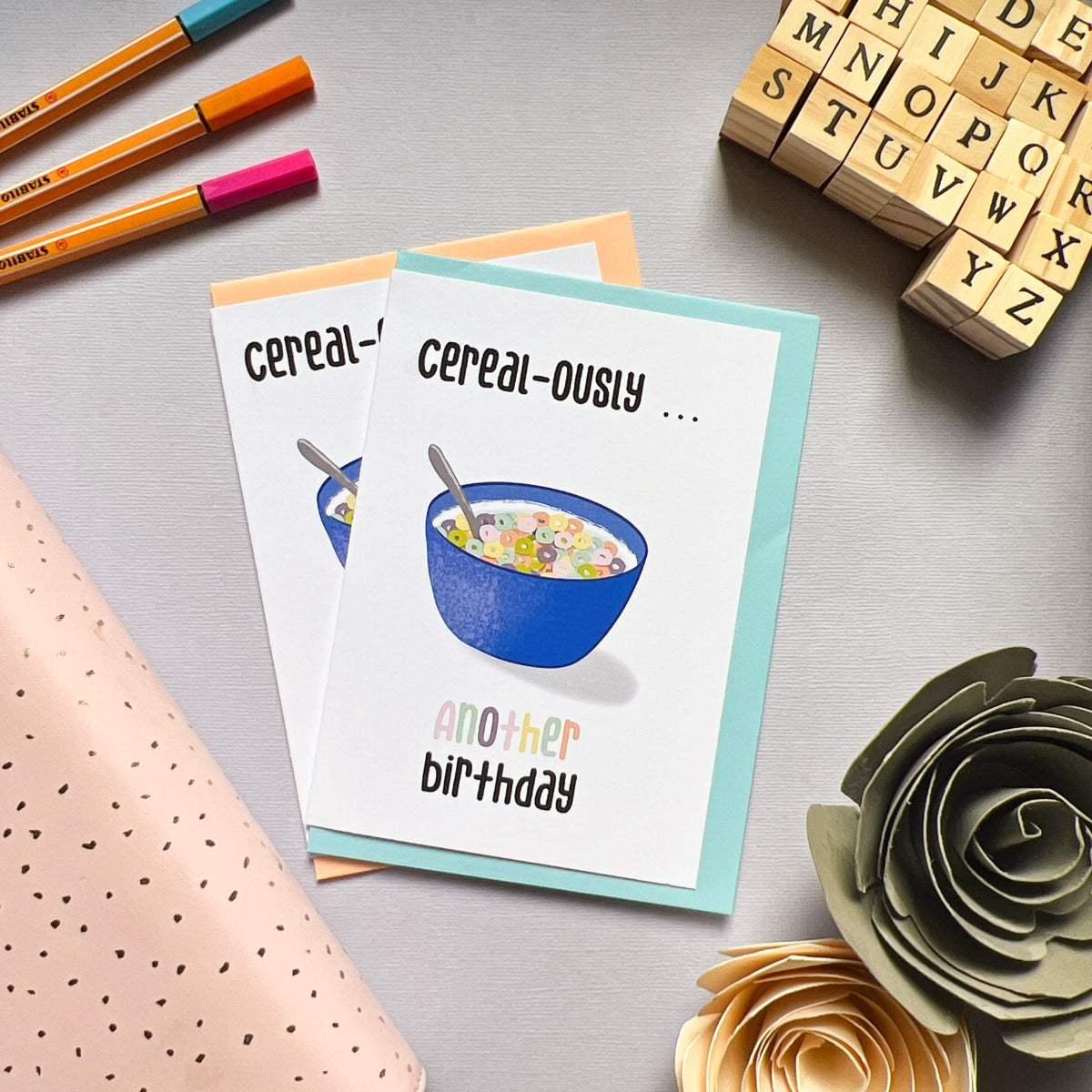 Two "Cereal-ously Another Birthday" greeting cards placed on a table each feature an illustration of a blue bowl filled with colorful cereal and a spoon. The cards' covers display the words "cereal-ously... another birthday." Surrounding items include a letter stamp set, pens, and paper flowers. These cards are perfect for celebrating your friend's colorful birthday!