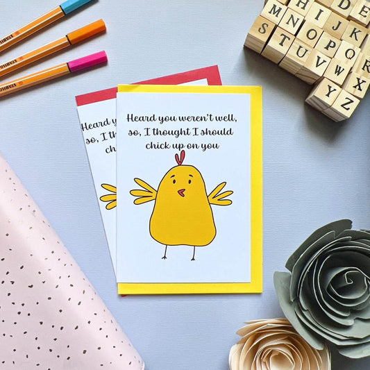 Two "Chicken up on you Get Well Soon" cards, featuring a cartoon chicken and the text "Heard you weren't well, so, I thought I should chick up on you," are displayed on a flat surface. Surrounding the cards are brightly colored envelopes, markers, a wrapped gift, wooden alphabet stamps, and decorative paper flowers.