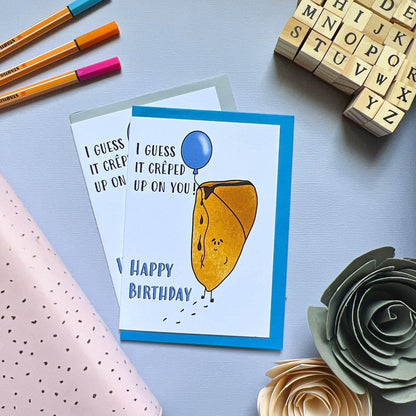 Product Name: Birthday Crepe Card

A crêpe card featuring an illustration of a crepe holding a blue balloon, accompanied by the text "I guess it creped up on you! Happy Birthday." The card is placed on a table with colorful pens, alphabet stamps, and decorative paper flowers nearby. It's presented in biodegradable packaging.