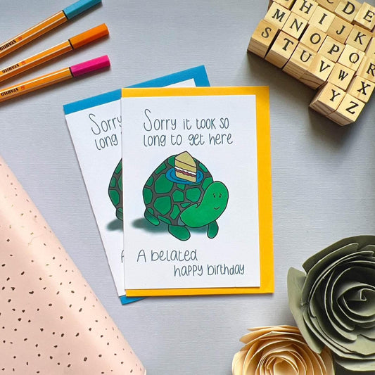 Two "Belated Birthday Cards" feature a turtle carrying a slice of cake on its shell. The message reads, "Sorry it took so long to get here. A belated happy birthday." The cards are placed on a gray surface next to colorful pens, large decorative paper flowers, and wooden letter stamps.