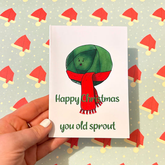 A hand presents a Christmas Sprout Blank Christmas Card showcasing a digitally illustrated sprout adorned in a red scarf. The card's message reads, "Happy Christmas you old sprout," set against a lively backdrop of festive holiday hats.