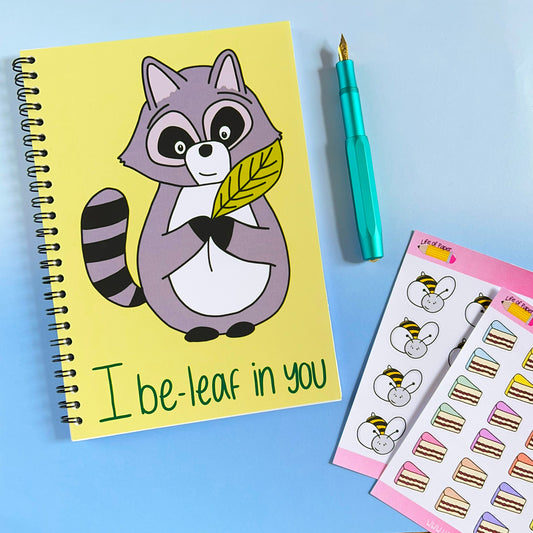 An I Be-leaf In You Notebook with a cartoon raccoon holding a leaf and the text "I be-leaf in you" sits beside a teal fountain pen and two sheets of stickers featuring cartoon animals and pastel-colored cakes. This encouraging notebook is set against a blue background.