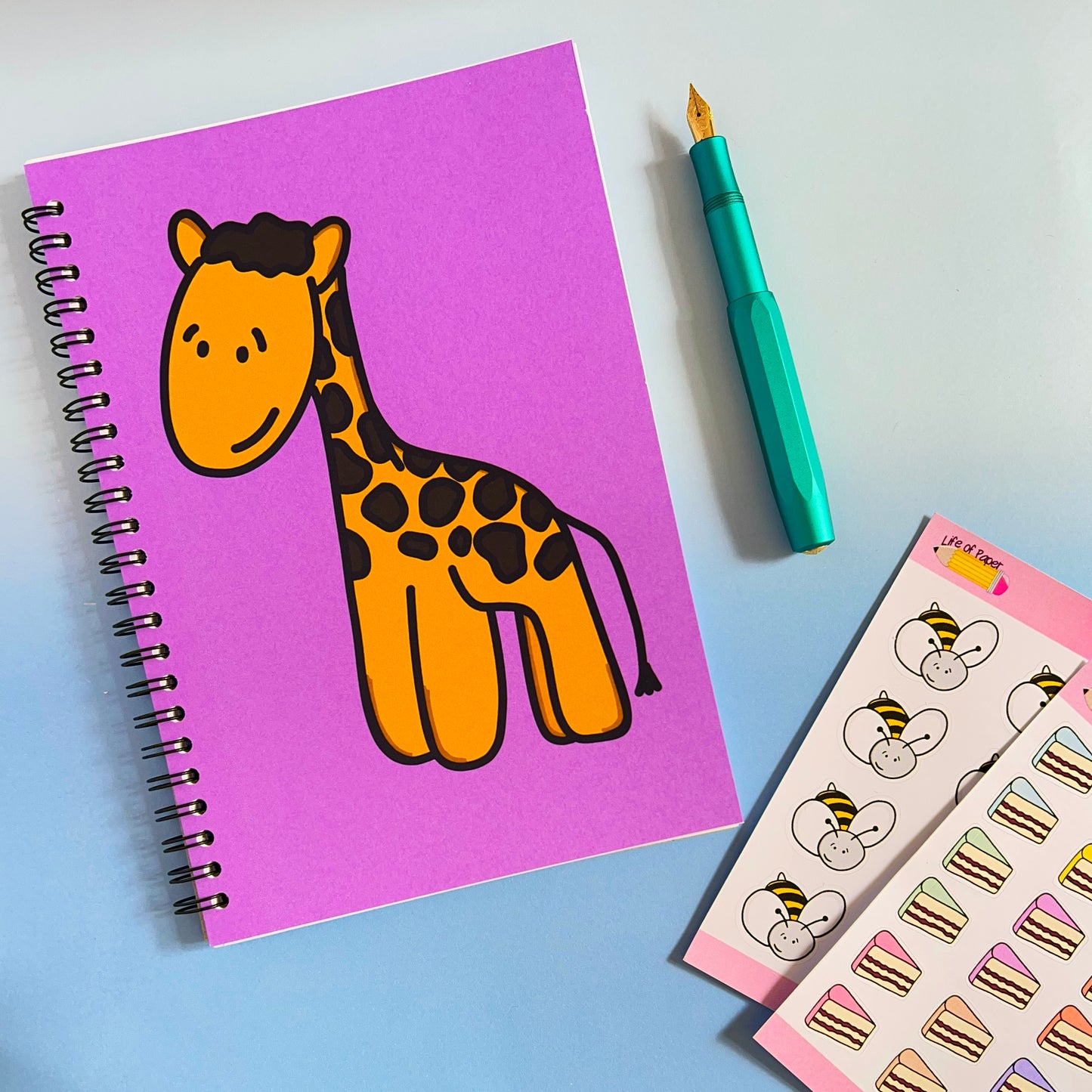 A vibrant workspace featuring a Happy Giraffe Notebook with a soft pink cover, a teal fountain pen, and a sheet of cute stickers with animal faces and cakes against a light blue background. The notebook includes 40 pages, perfect for jotting down your thoughts.