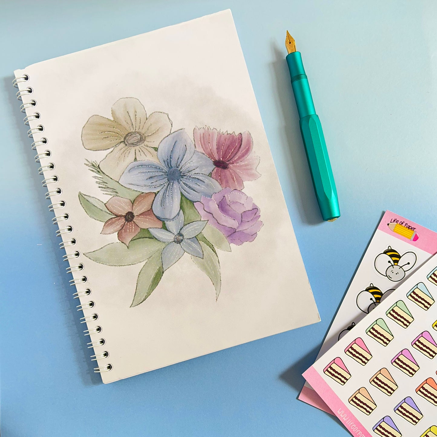 A sketchbook with a drawing of colorful flowers rests on a blue surface. Beside the sketchbook is a teal fountain pen and a sheet of stickers featuring cake slices and cartoon faces, including bears and bees. This high-quality Faded Floral Notebook adds charm to any artist's collection.