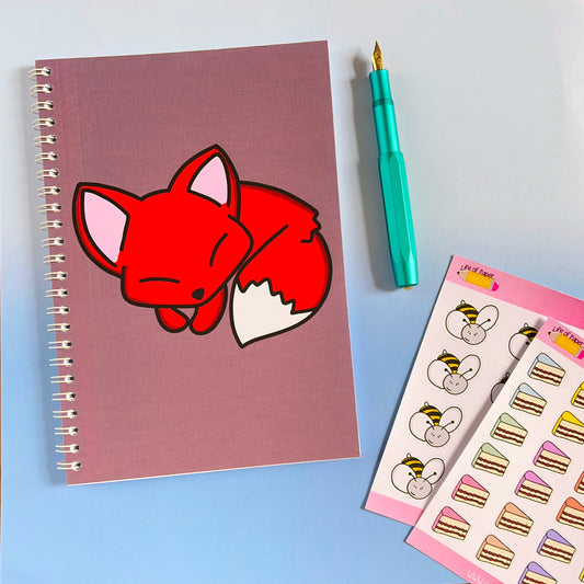The 40-page "Sleepy Fox Notebook" features a red, sleeping fox illustration on its purple spiral-bound cover. It's accompanied by a light blue fountain pen and a sheet of colorful stickers with animal faces and small cake slices, set against an autumnal toned gradient background of light blue and white.