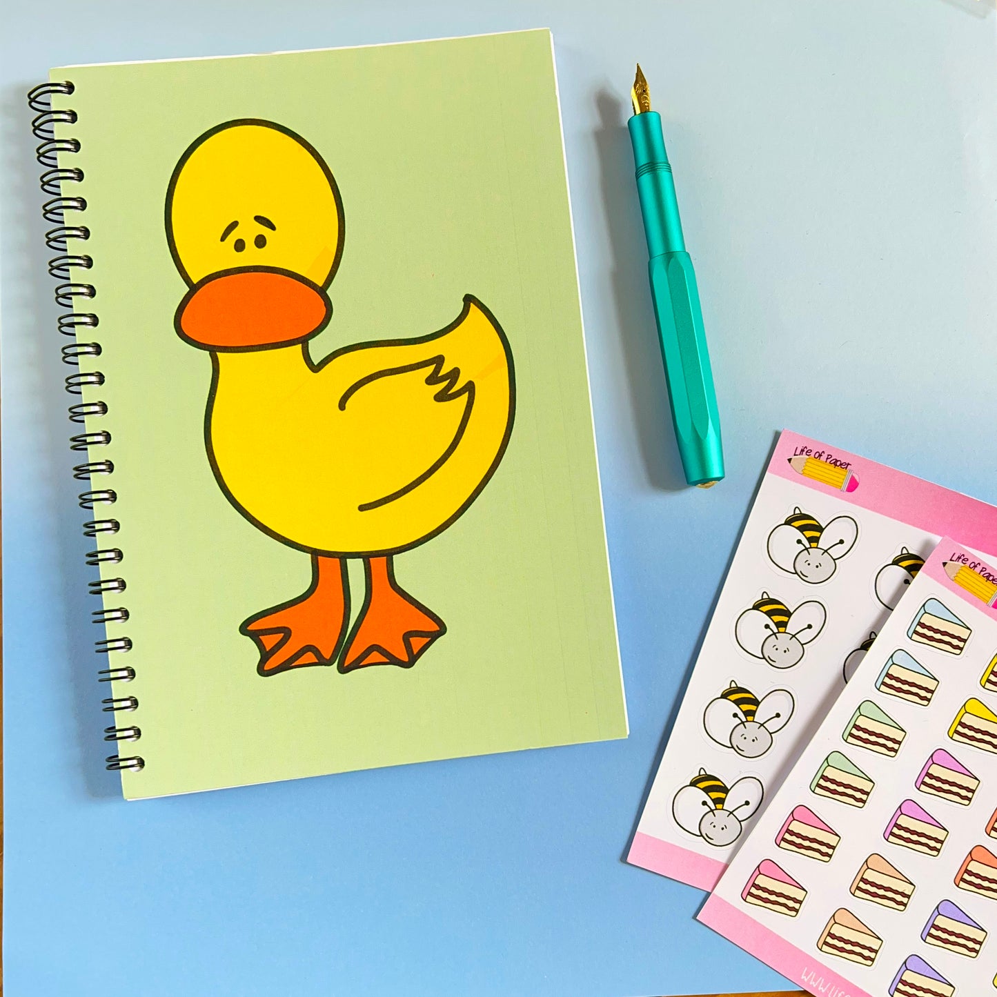 A Worried Duck Notebook with a cartoon duck on the cover is placed on a light blue surface. To the right of the notebook is a teal fountain pen and two sheets of colorful stickers featuring animal faces and layered cakes, perfect for note-taking.