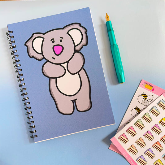 A blue spiral-bound Gentle Koala Notebook with 40 pages sits on a pastel surface. Next to it, a teal fountain pen and a sheet of stickers featuring cake slices and a cartoon bear await your note-taking delight.