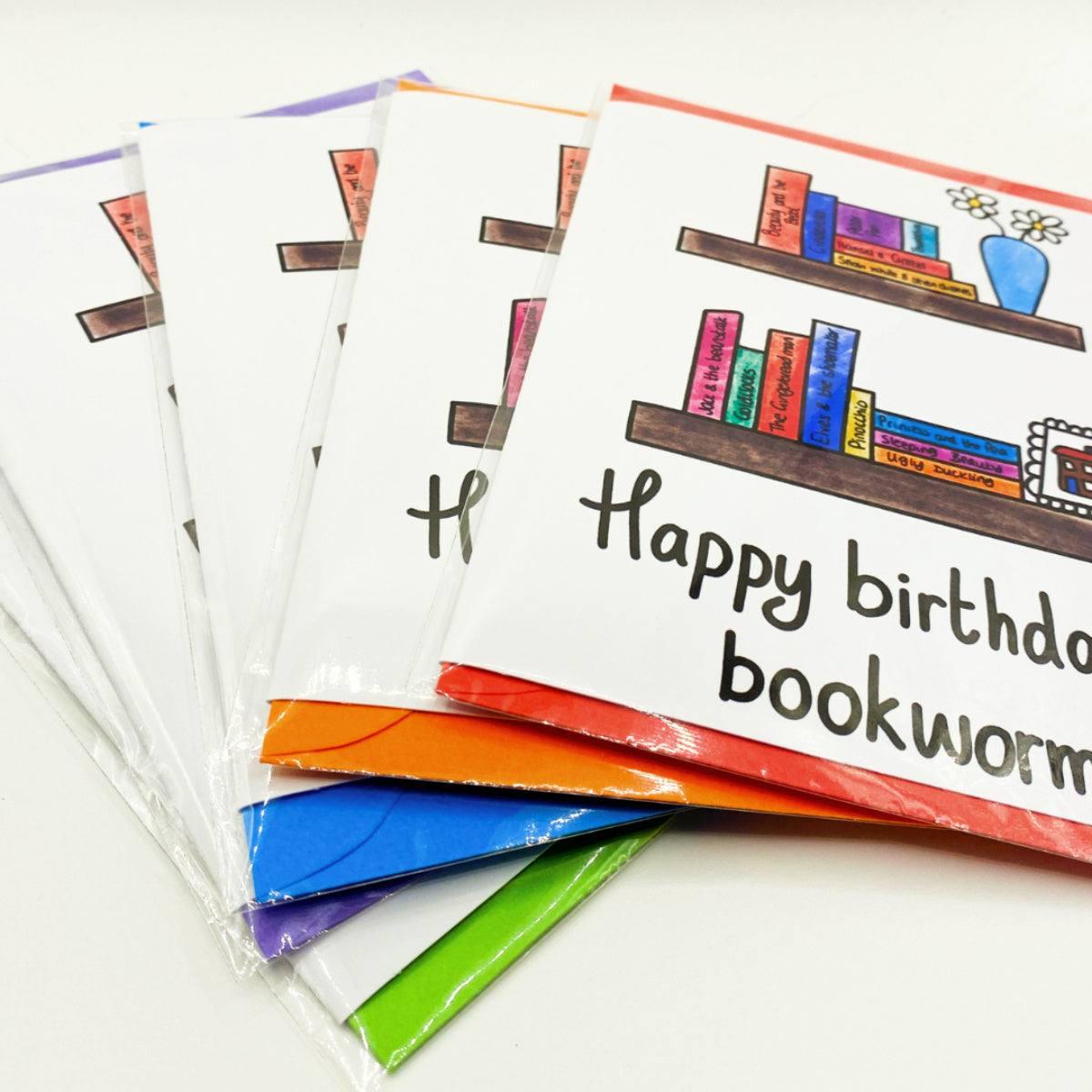 A stack of the Bookworm Birthday Card features a hand-drawn bookshelf adorned with books and a potted plant. The front of each birthday card proudly displays the message "Happy birthday, bookworm!" Every card is enclosed in a clear plastic sleeve.