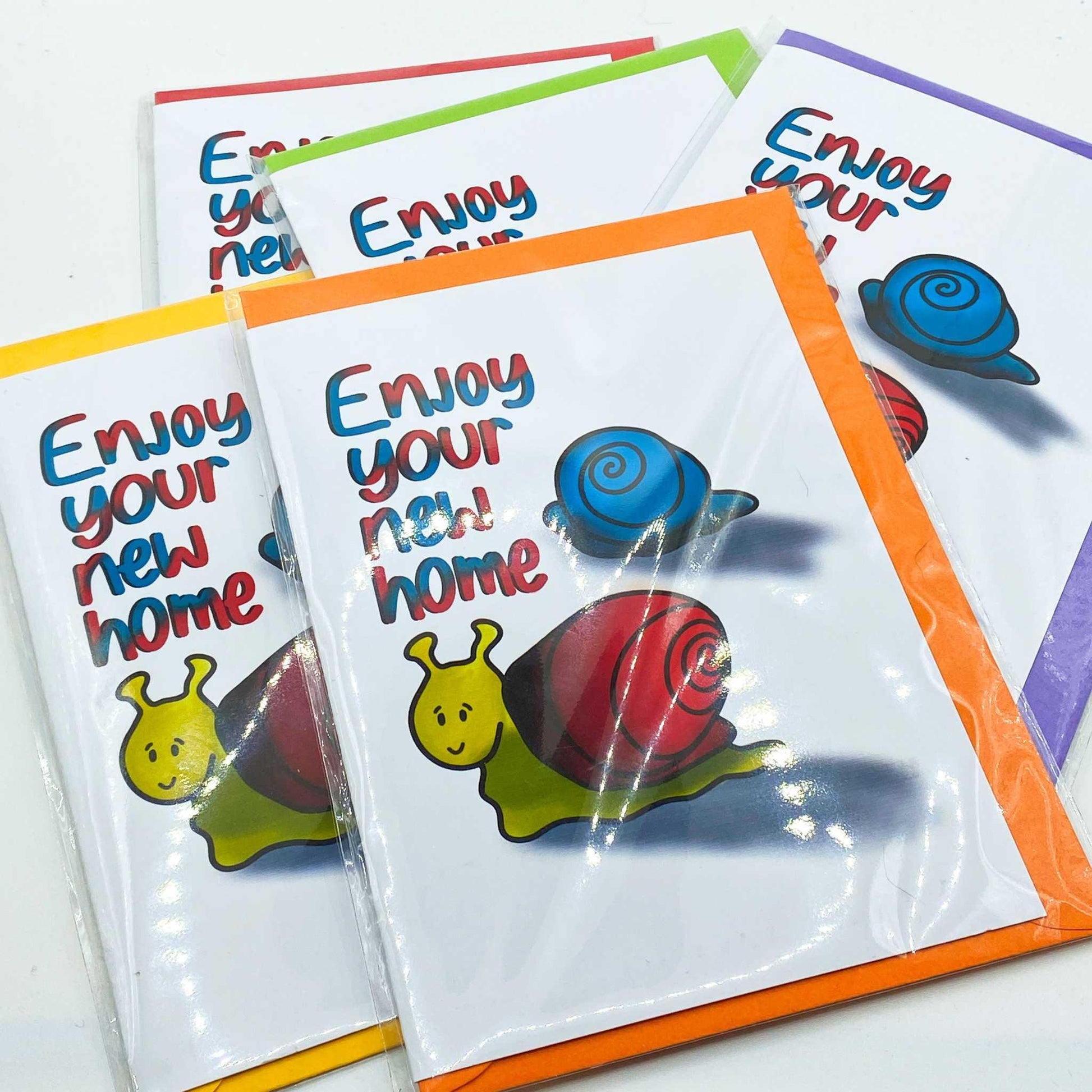 A display of five New Home Greeting Cards showcases colorful borders and features illustrated snails alongside the text "Enjoy your new home." Each card presents a smiling yellow snail with a red shell in the foreground and a blue snail in the background, all encapsulated in biodegradable packaging.