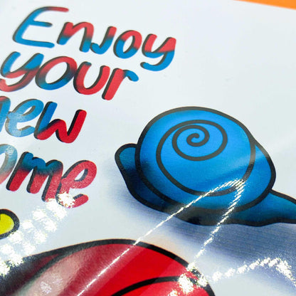 The New Home Greeting Card is a glossy card featuring colorful text that reads "Enjoy your new home" in a playful font. On the right side, there's an illustration of a blue snail with a spiral shell, adding a whimsical touch to this charming card. The thoughtful design comes in biodegradable packaging.