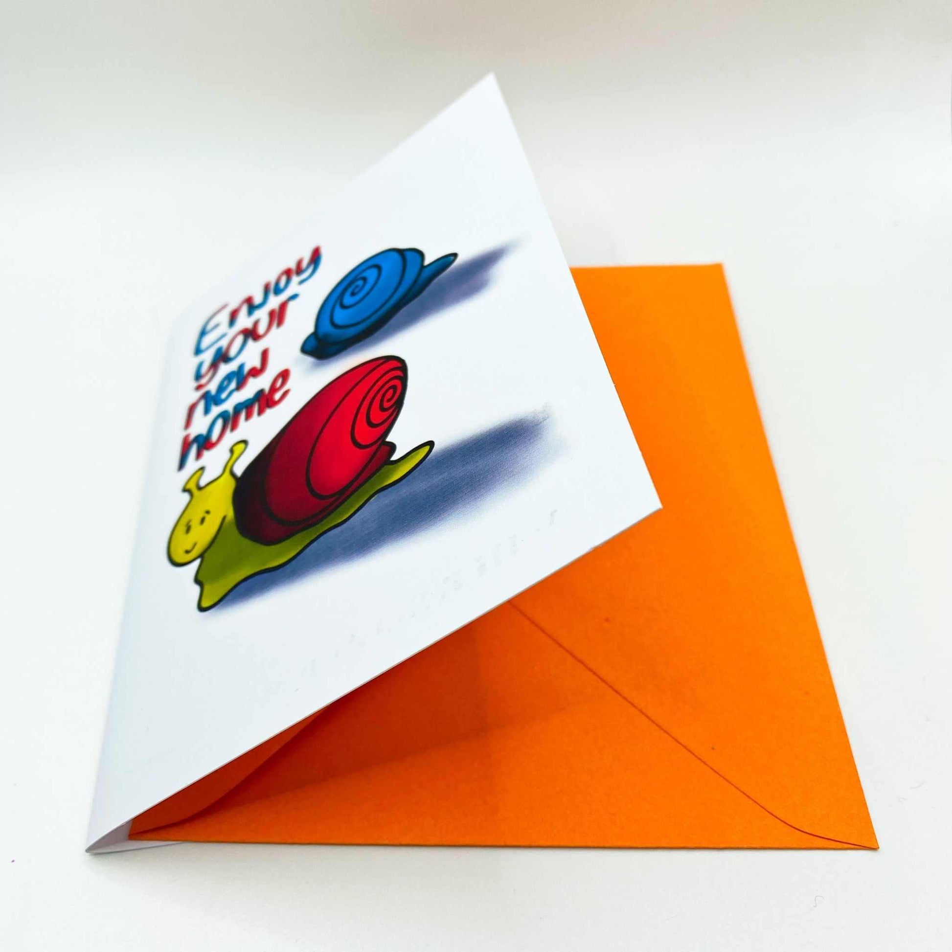 Introducing the New Home Greeting Card: This delightful card showcases a yellow snail with a red shell and a blue snail with a blue shell, adorned with the text "Enjoy your new home." It comes partially open in an orange envelope, adding to its charm. Packaged in biodegradable materials, this eco-friendly greeting card is perfect for celebrating a new beginning.