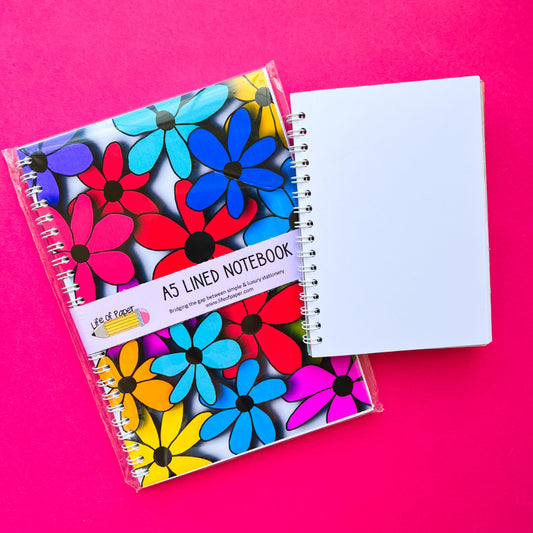 Two spiral notebooks on a pink background: one is the A6 Big Floral Notebook, a high-quality notebook adorned with a vibrant floral cover and labeled "You Are Enough," while the other features a simple, plain white design.