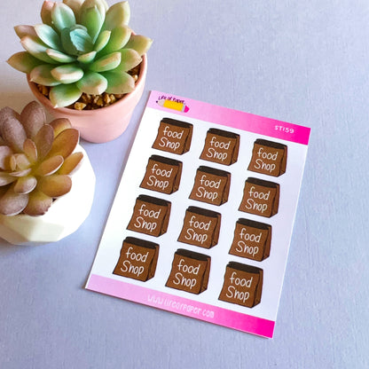 A sheet of stickers labeled "Food Shop Planner Stickers" is displayed on a flat surface between two small potted succulents. The sheet, perfect for weekly planning, has a pink header with product information and contains nine brown rectangular planner stickers with the text "food shop.
