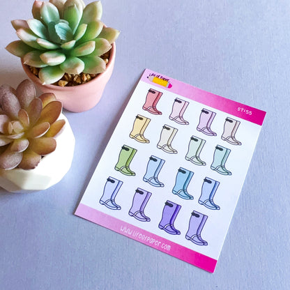 A sheet of colorful Wellington Boot Planner Stickers is placed on a purple surface. Next to the sheet are two small succulent plants in white and pink pots. The top edge of the rainy day sticker sheet is pink, displaying the brand's logo and website URL.