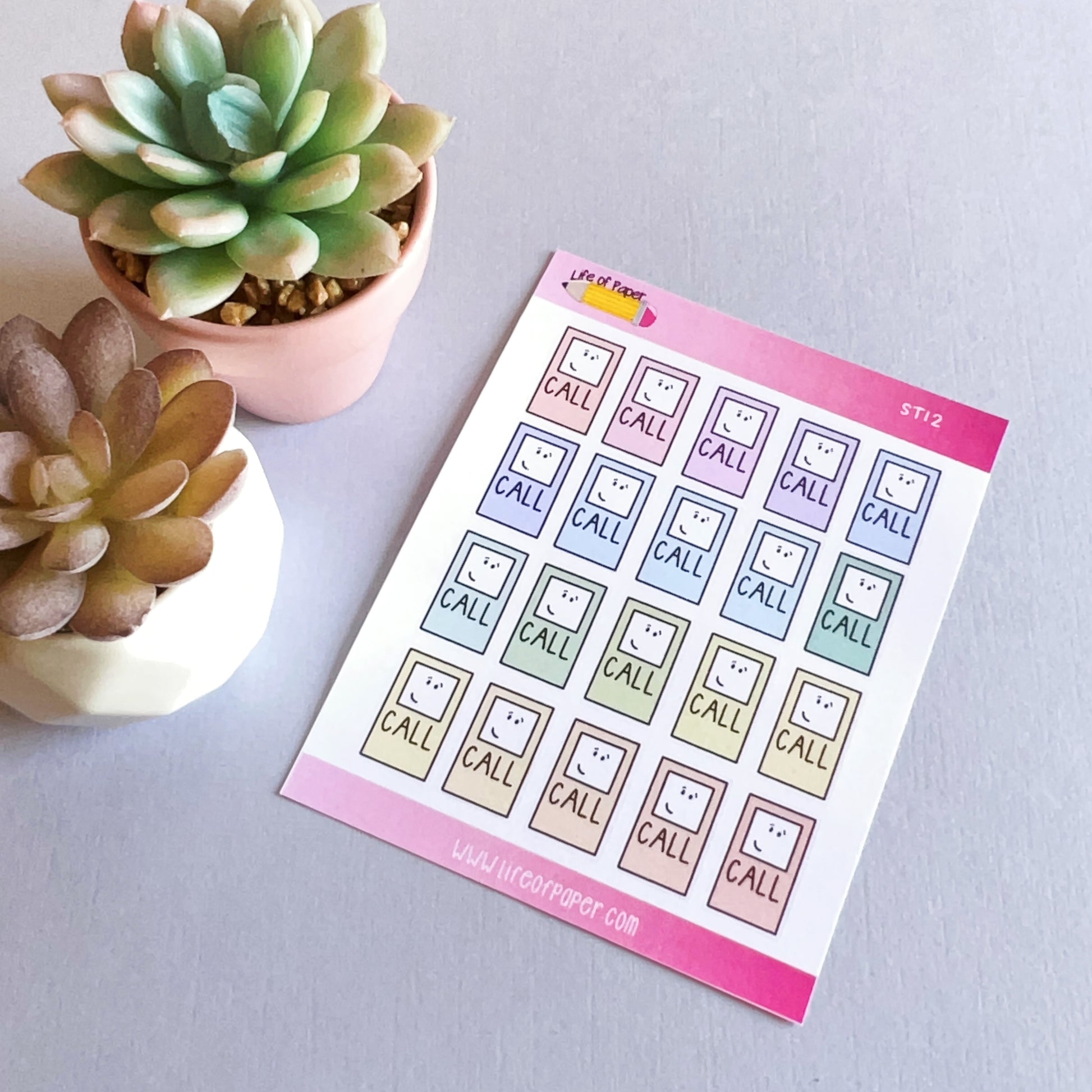 A sheet of colorful Call Reminder Planner Stickers featuring a smiling phone icon above the word "CALL" is displayed on a light grey surface. Next to the sheet, there are two potted succulents in white and pink pots, making it perfect for adding some life to your planner or diary.