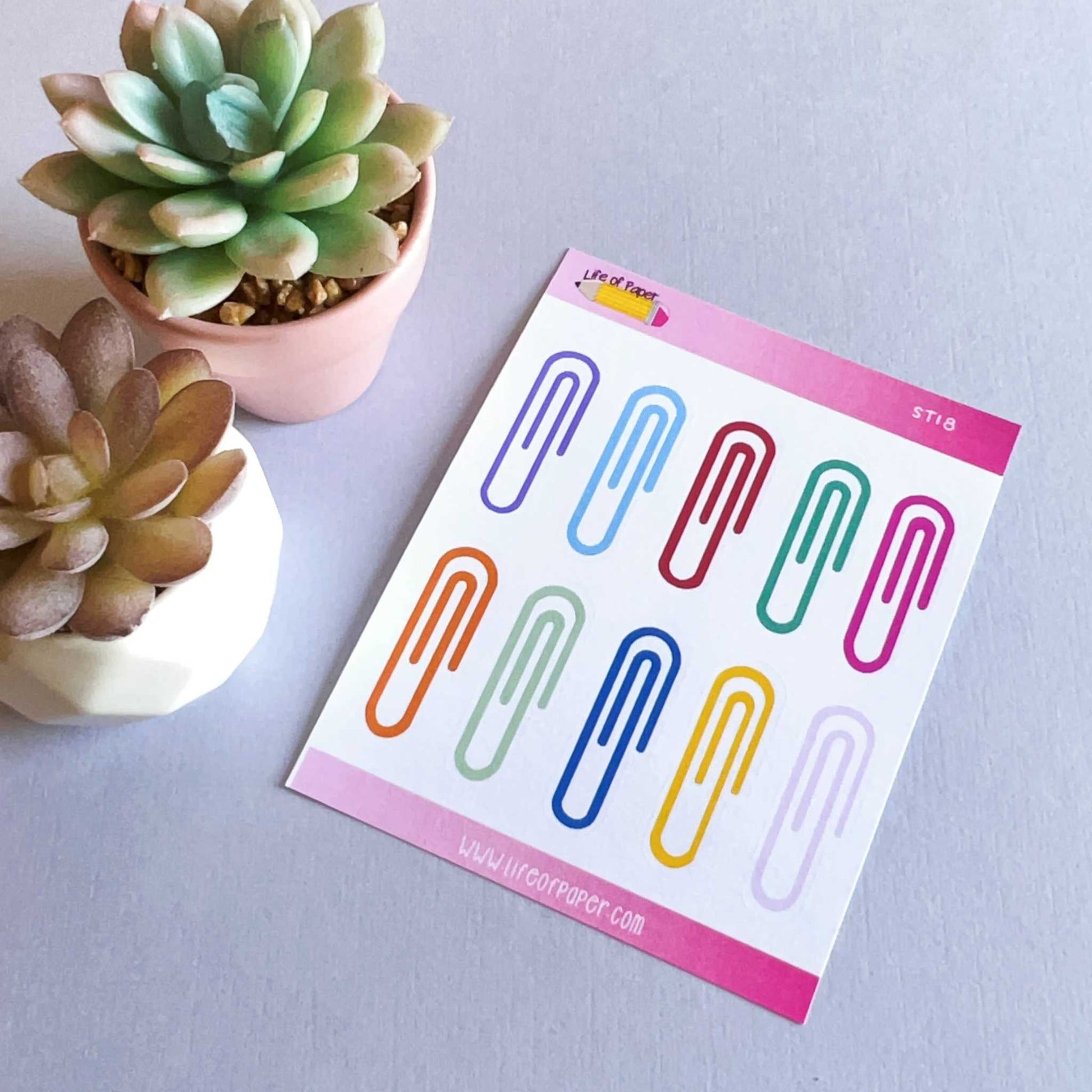 A pink and white sheet with Rainbow Paperclip Planner Stickers sits on a gray surface next to two small potted succulent plants, one in a round white pot and the other in a beige pot.