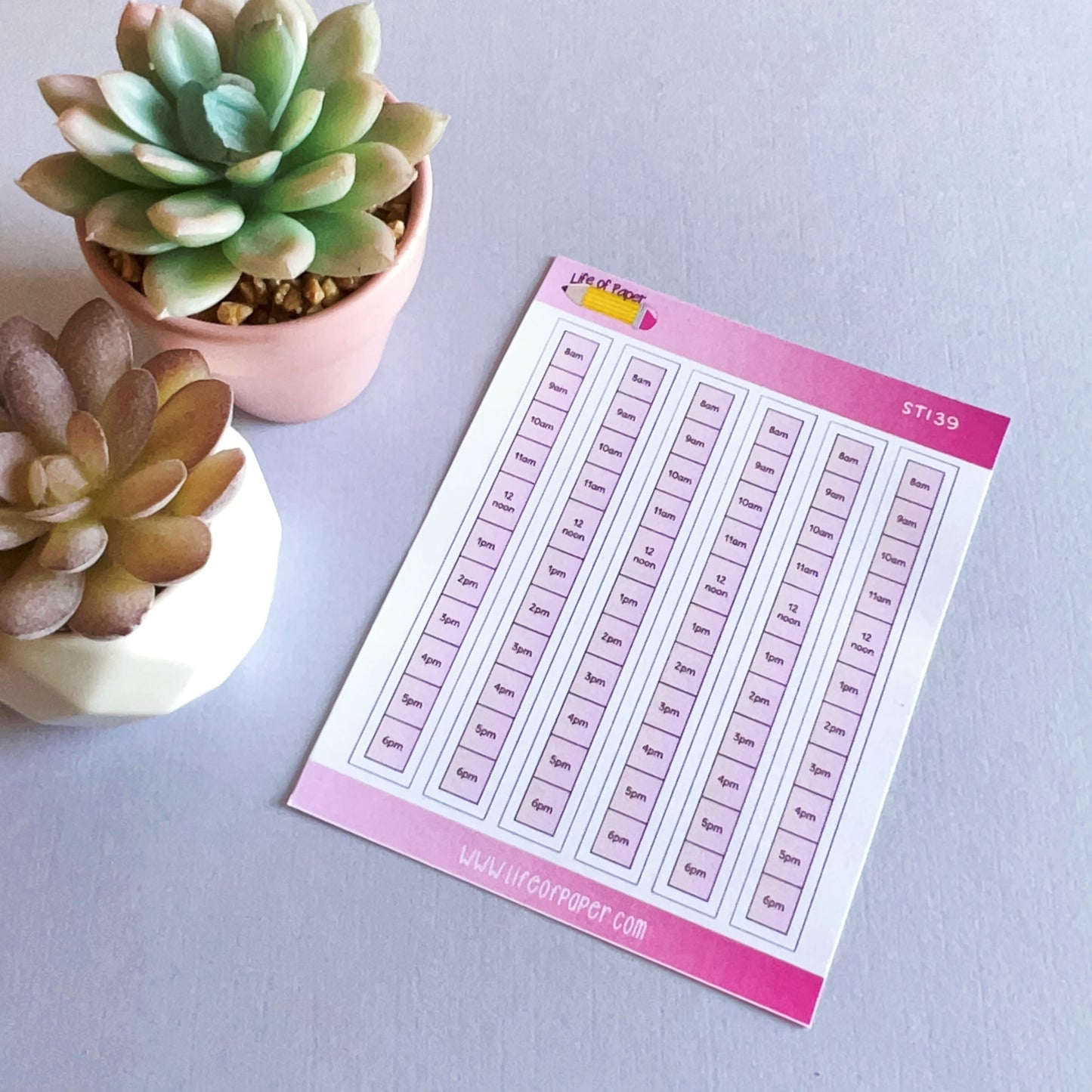 A sheet of Daily Schedule Planner Stickers labeled with times from 7am to 6pm is placed on a flat surface. Two succulent plants in small pots, one white and one pink, are positioned to the left of the sticker sheet, ready to enhance your daily schedule. The background is a light grey color.
