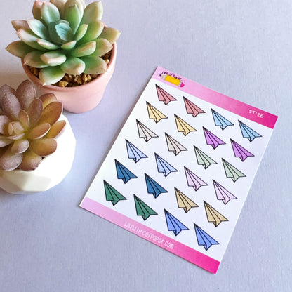 A Paper Aeroplane Sticker Sheet lies on a light surface, accompanied by two small succulent plants in white pots. The stickers are arranged in neat rows, featuring various colors including blue, yellow, green, pink, and purple.