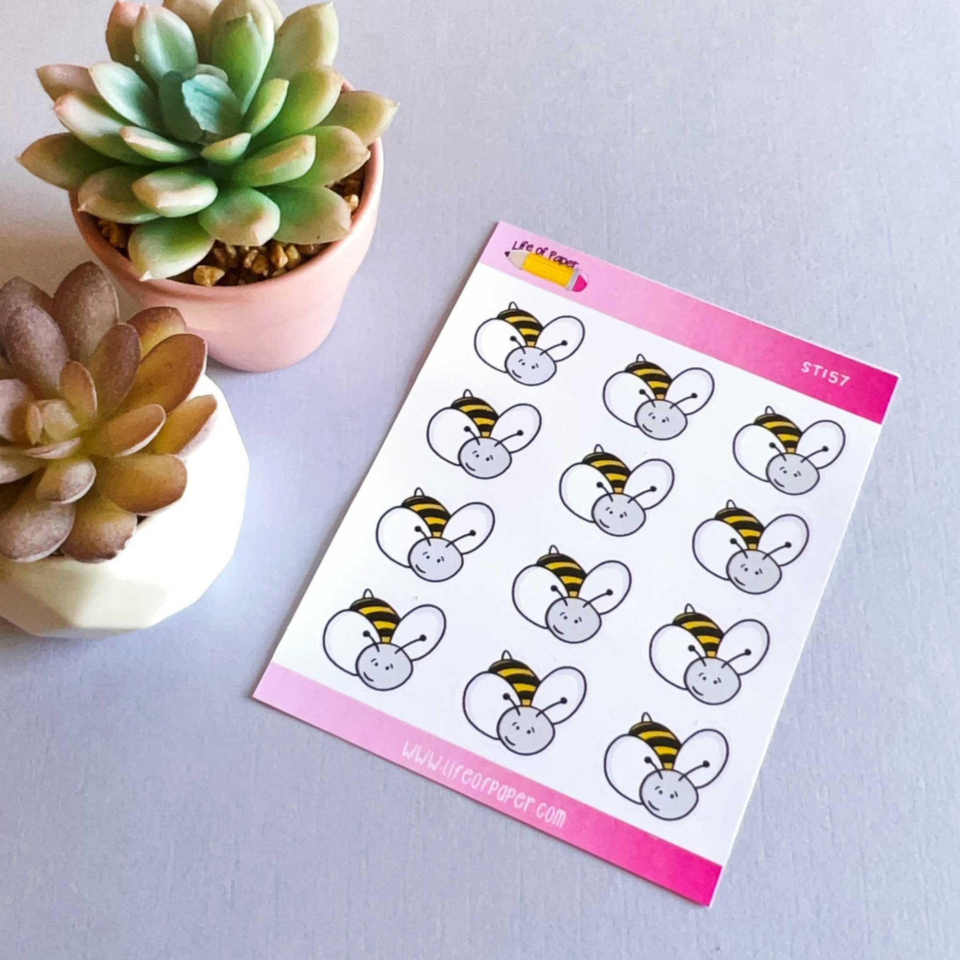 A Bumblebee Sticker Sheet featuring a cartoon bunny in a bee costume is placed on a flat surface. Next to it, two small succulent plants in pots, one green and one multicolored, add a touch of nature. The background remains a simple light grey.