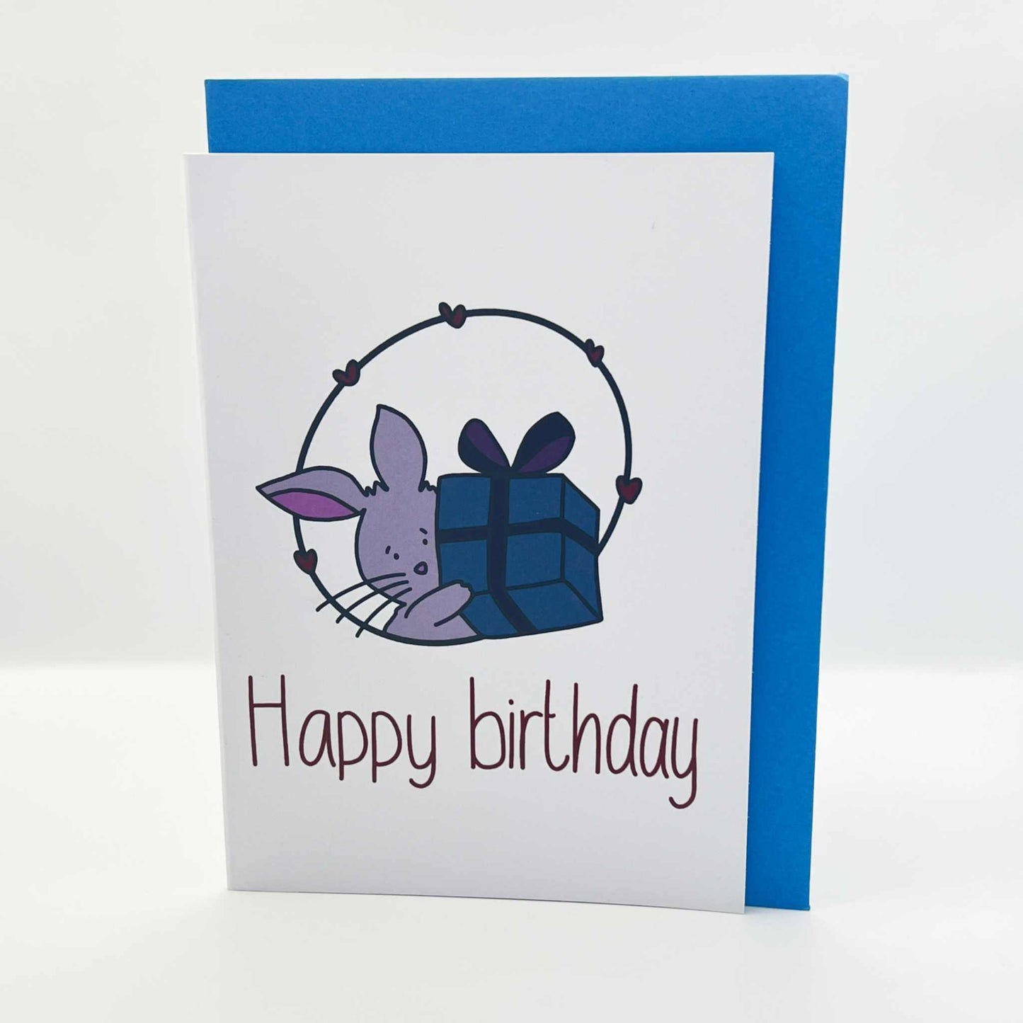 Introducing the Cute Bunny Birthday Card, featuring an adorable bunny peeking from behind a wrapped gift topped with a bow. The card showcases a white front with "Happy Birthday" elegantly printed in purple and comes paired with a charming blue envelope.