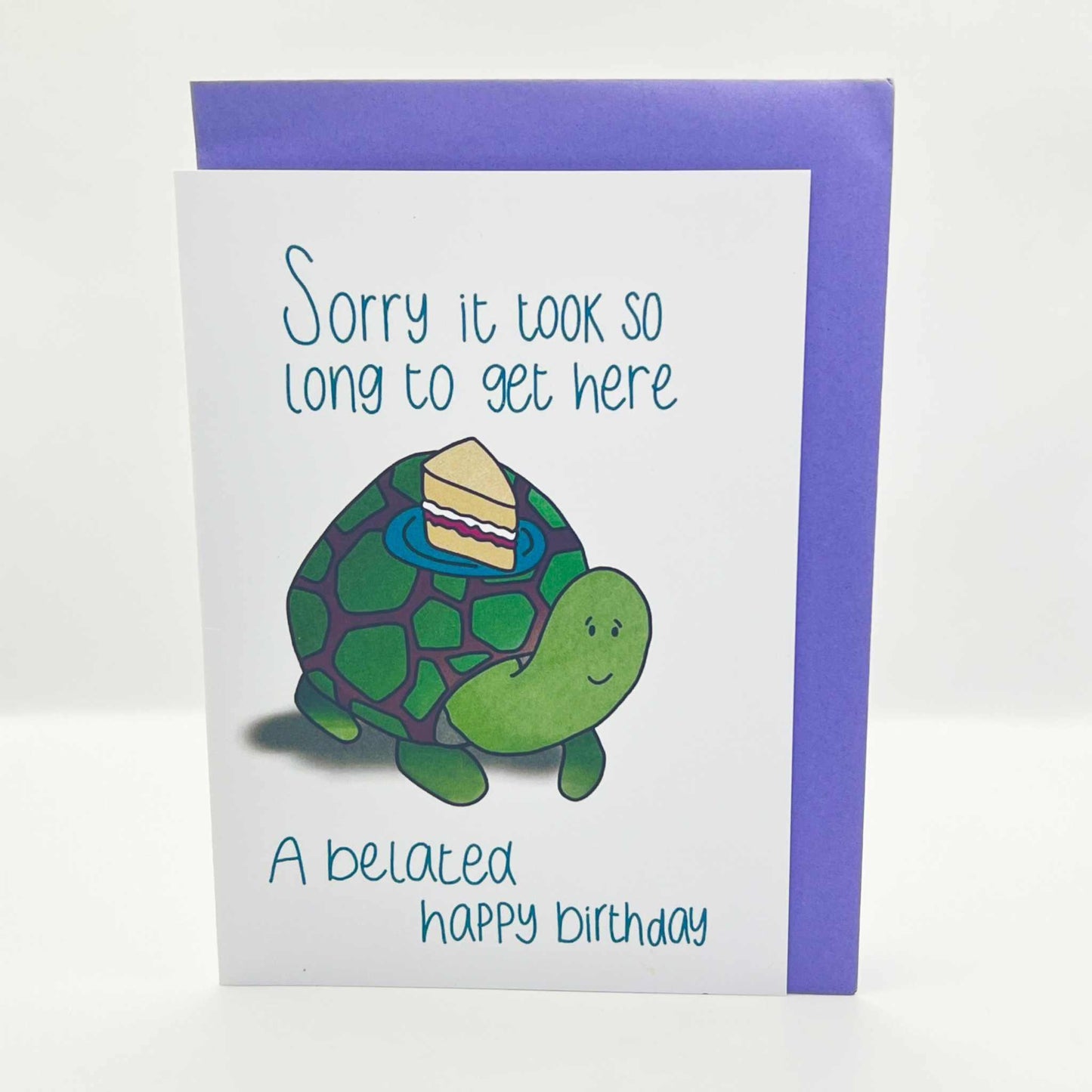 The Belated Birthday Card is a biodegradable card featuring a charming drawing of a smiling turtle with a slice of cake on its shell. The text reads, "Sorry it took so long to get here. A belated happy birthday." The card is bordered in purple and set against a plain white background.