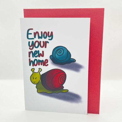 The New Home Greeting Card displays two charming illustrated snails, one blue and one green with a red shell, accompanied by bright and playful lettering that reads "Enjoy your new home." It comes paired with a vibrant red envelope.