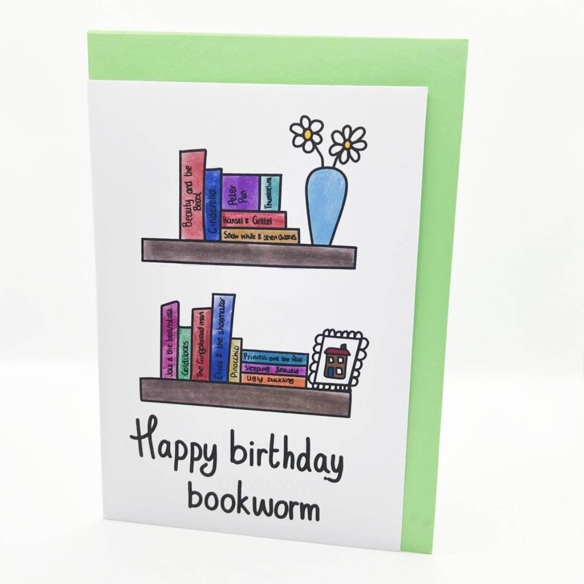 The Bookworm Birthday Card boasts a light green background with an illustration of two bookshelves. The top shelf displays colorful books and a blue vase containing two daisies, while the bottom shelf holds additional books and a small framed picture. The card features the message "Happy birthday bookworm!" making it an ideal gift for any book lover!