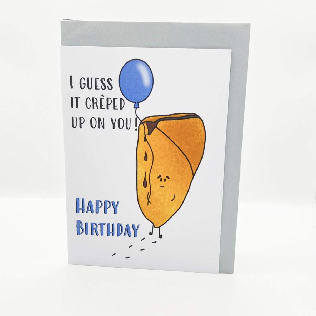 The Birthday Crepe Card displays the message "I guess it crêped up on you! Happy Birthday" on the front, featuring an illustration of a crêpe with a face and legs, holding a blue balloon. The whimsical crêpe appears to be walking and leaving small marks behind. This charming card is also eco-friendly, arriving in biodegradable packaging.