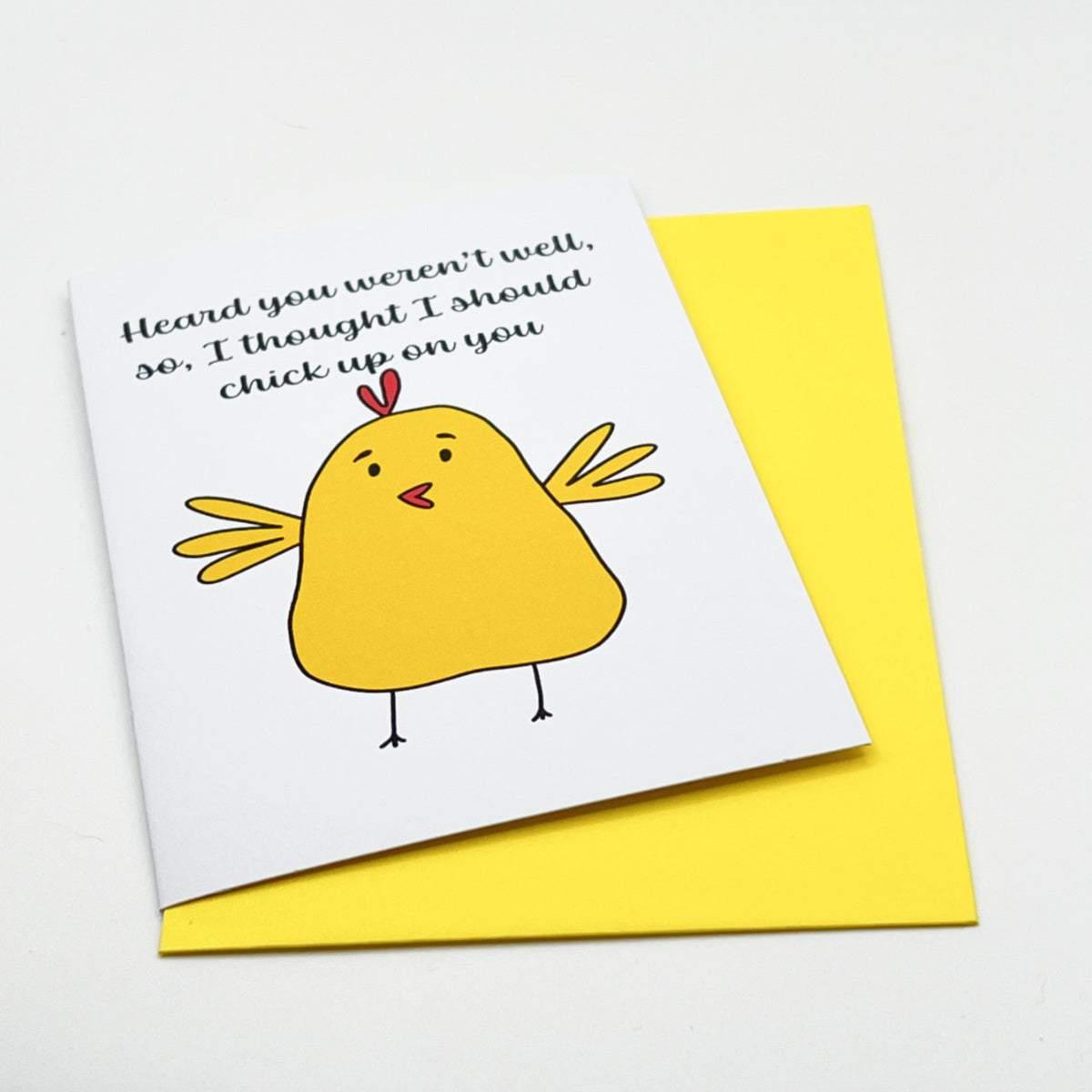 The "Chicken up on you Get Well Soon Card" showcases a charming cartoon yellow chick with its wings spread wide. The card has the message, "Heard you weren't well, so I thought I should chick up on you," and comes with a brightly colored envelope. It is enclosed in biodegradable cellophane and presented against a white background.