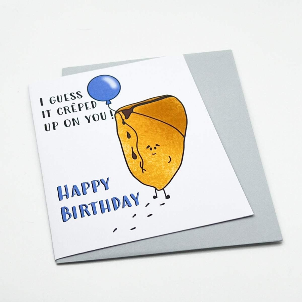 The Birthday Crepe Card features a charming illustration of a crepe holding a blue balloon and looking surprised. The card reads, "I guess it crêped up on you! Happy Birthday." Propped up against a plain, light grey envelope on a white surface, it comes in biodegradable packaging.