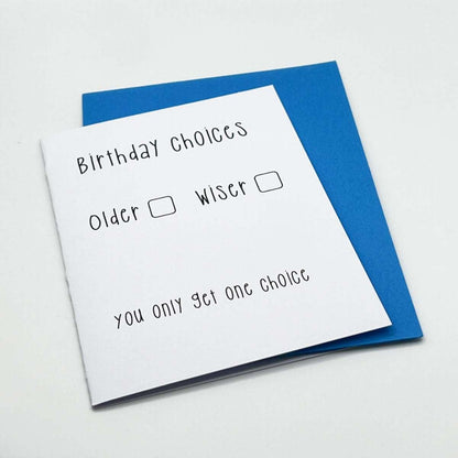 The Birthday Choices Blank Card features a white background with black text that reads, "Birthday Choices: Older [ ] Wiser [ ]. You only get one choice." Perfect for celebrating an older friend's special day, the card is showcased on a blue envelope against a white surface. Made from eco-friendly and biodegradable materials, this card adds a thoughtful touch to any celebration.