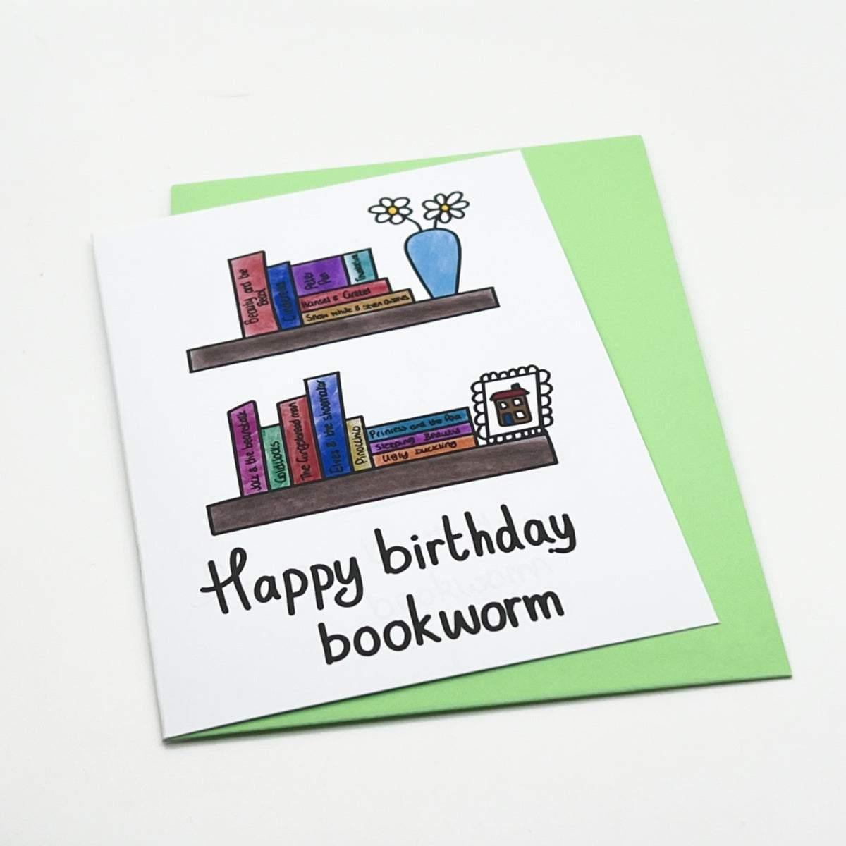 The Bookworm Birthday Card comes with a green envelope and showcases vibrant illustrations of two bookshelves. The top shelf displays an array of books alongside a blue vase filled with flowers, while the bottom shelf holds more books and a picture frame. This playful design is completed with the handwritten message "Happy birthday bookworm.