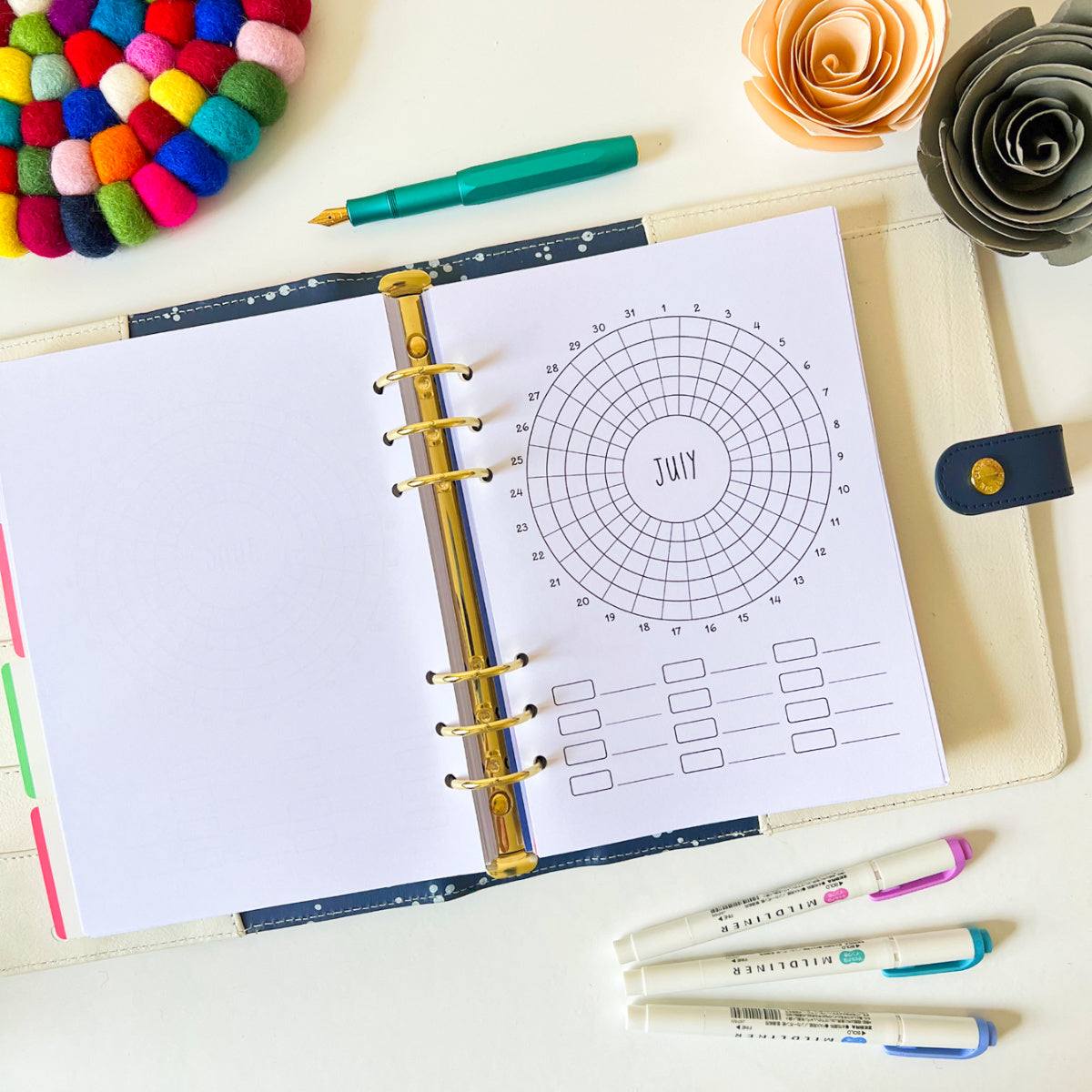 An open Monthly Habit Tracker lies on a table, displaying a circular July calendar spread and a section for tasks and goal setting. Surrounding the tracker are colorful markers, a green pen, and decorative items including a multicolored pom-pom coaster and paper flowers.