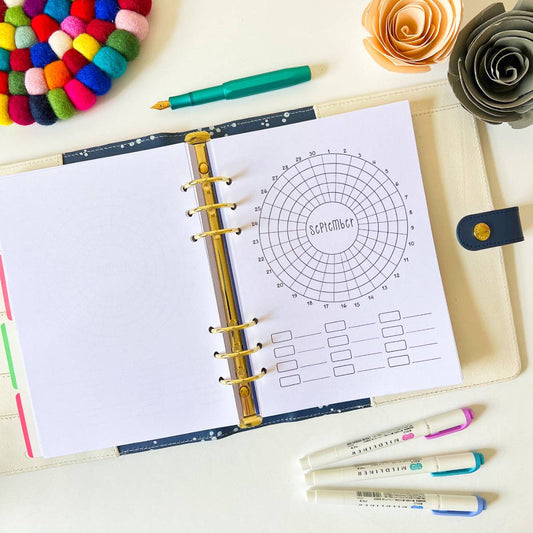A planner lies open on a desk with a circular calendar for September vertically aligned in the center. Surrounding it are colorful accessories including a pink and green pen, a rainbow pom-pom coaster, decorative paper flowers, and a Monthly Habit Tracker to aid in goal setting.
