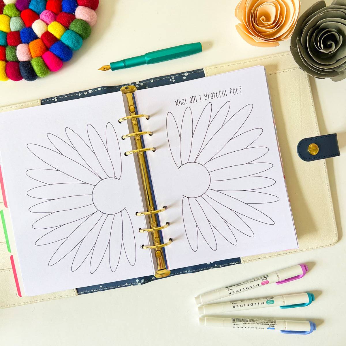 A Monthly Gratitude Log open to a page with two large, blank daisy illustrations to be filled in. Above the daisies, the words "What am I grateful for?" are printed. Surrounding the log are a colorful coaster, pens, a closed memory planner, and a paper flower.