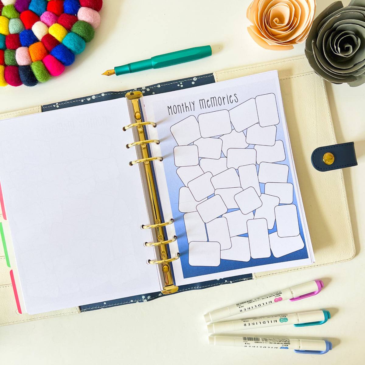 A planner lies open on a table, displaying a "Memory Log" page perfect for capturing positive moments. Surrounding the planner are colorful markers, a teal pen, a wool coaster, and two decorative paper flowers, inviting you to log everyday life's highlights.
