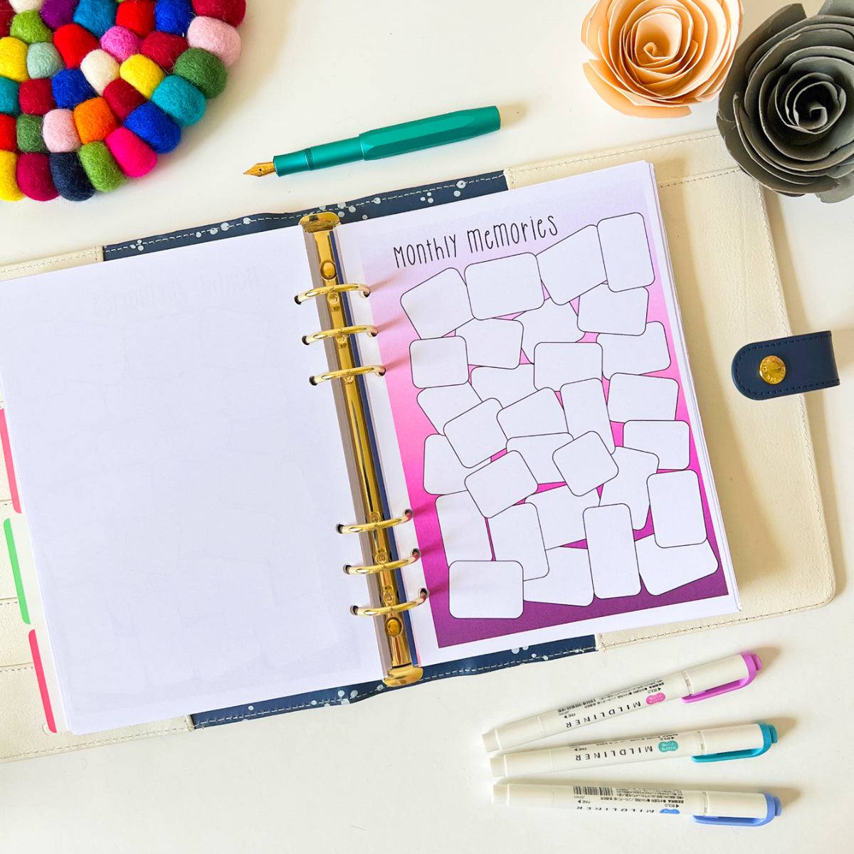 An open planner lies on a table with a "Memory Log" page displaying blank boxes for notes, perfect for capturing positive moments of everyday life. Nearby are colorful markers, a green pen, two decorative flower figures, and a multicolored pom-pom coaster, creating a vibrant workspace.