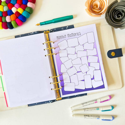 A planner lies open on a white desk, displaying a "Memory Log" page filled with blank rectangle shapes ready to log positive moments. Nearby, colorful pens, a green fountain pen, a multicolored pom-pom coaster, and two decorative paper flowers are arranged neatly.