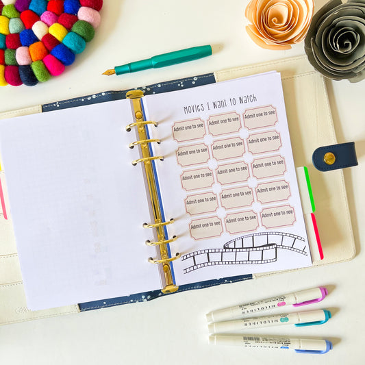 An open planner page is displayed on a white surface with a section labeled "Movies I Want to Watch." The Film Tracker features ticket stub designs with "Admit one to see" text. Nearby are colorful pens, a wool felt ball coaster, and paper flowers arranged decoratively.